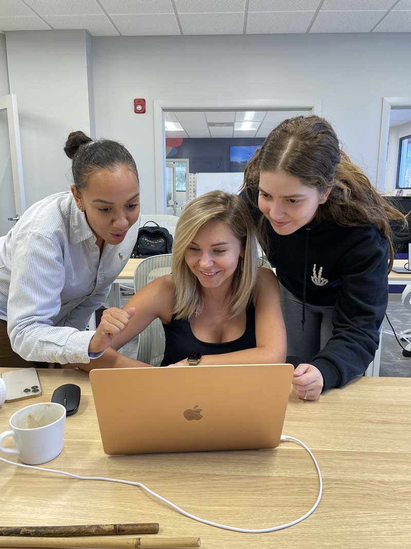 A diverse group of three young women collaborate and brainstorm over a single laptop.